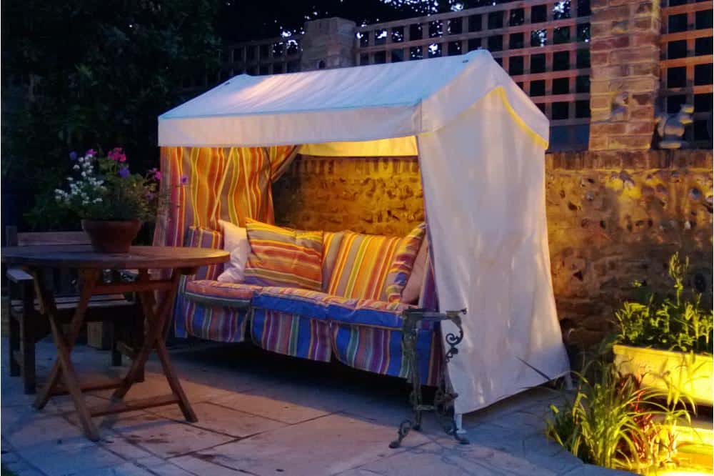 A cozy outdoor seating area features a cushioned bench with colorful striped pillows under a white canopy. The space is softly lit and includes a round wooden table with chairs nearby. A Wiverley's Idler swinging garden sofa adds charm, while stone walls and lattice fencing provide a rustic backdrop with greenery.