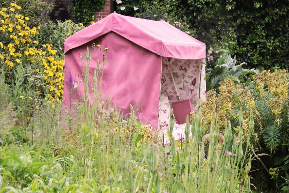 A small, pink fabric tent with floral-patterned sides stands in the middle of a lush, overgrown garden. Among the various green plants and yellow flowers creating a vibrant and natural setting, a Wiverley's Idler adds an inviting touch beside the tent.