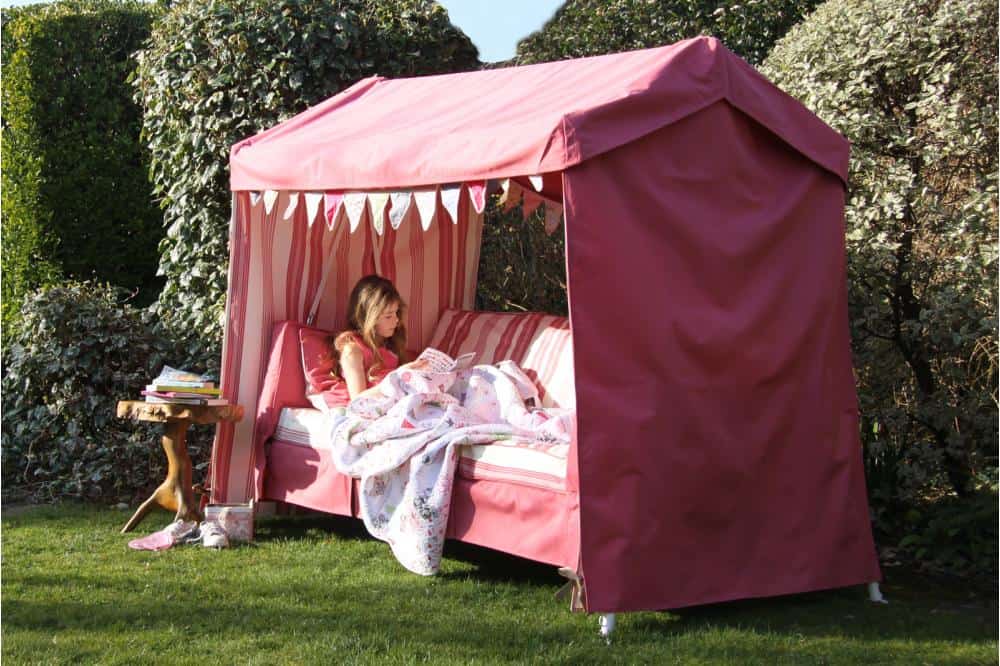 A young girl sits on a pink canopy bed with striped curtains, reading a book. The bed is set up outdoors on a lawn, near a Swinging Garden Sofa and surrounded by green bushes. A small wooden side table next to the bed holds a stack of Wiverley's Idler editions. The scene looks cozy and serene.