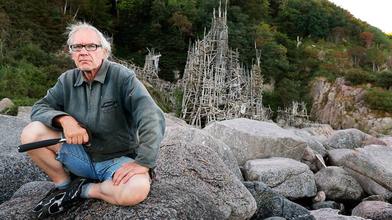 A man with glasses and a grey jacket sits on large rocks with a wood structure made of driftwood and a forested hill in the background. He grips a black object in his right hand, and his legs are exposed, wearing jean shorts. The scene appears to be outdoors in a rugged, natural environment.