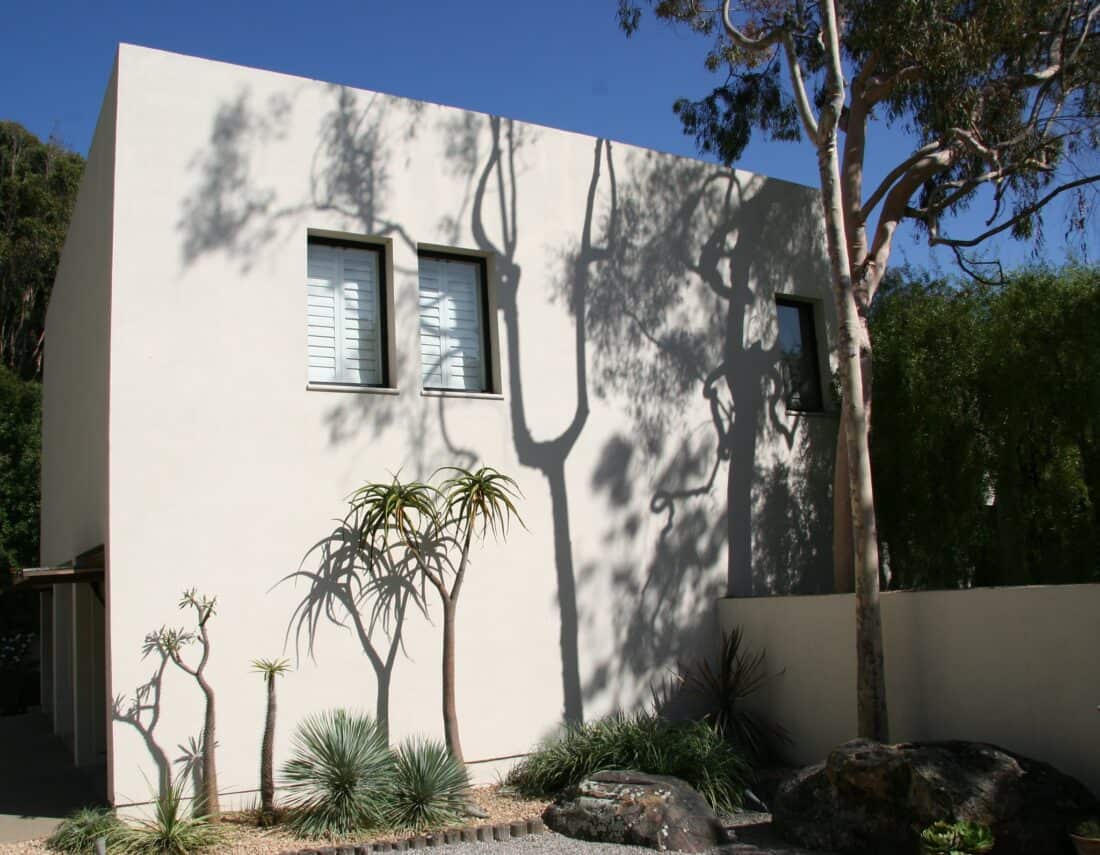 A minimalist two-story building with a smooth white facade and rectangular windows stands elegantly. Tall slender trees cast shadows on the wall, while Isabelle Greene's touch in the landscaping includes various desert plants and large rocks against a backdrop of greenery reminiscent of Santa Barbara. The sky is clear and blue.