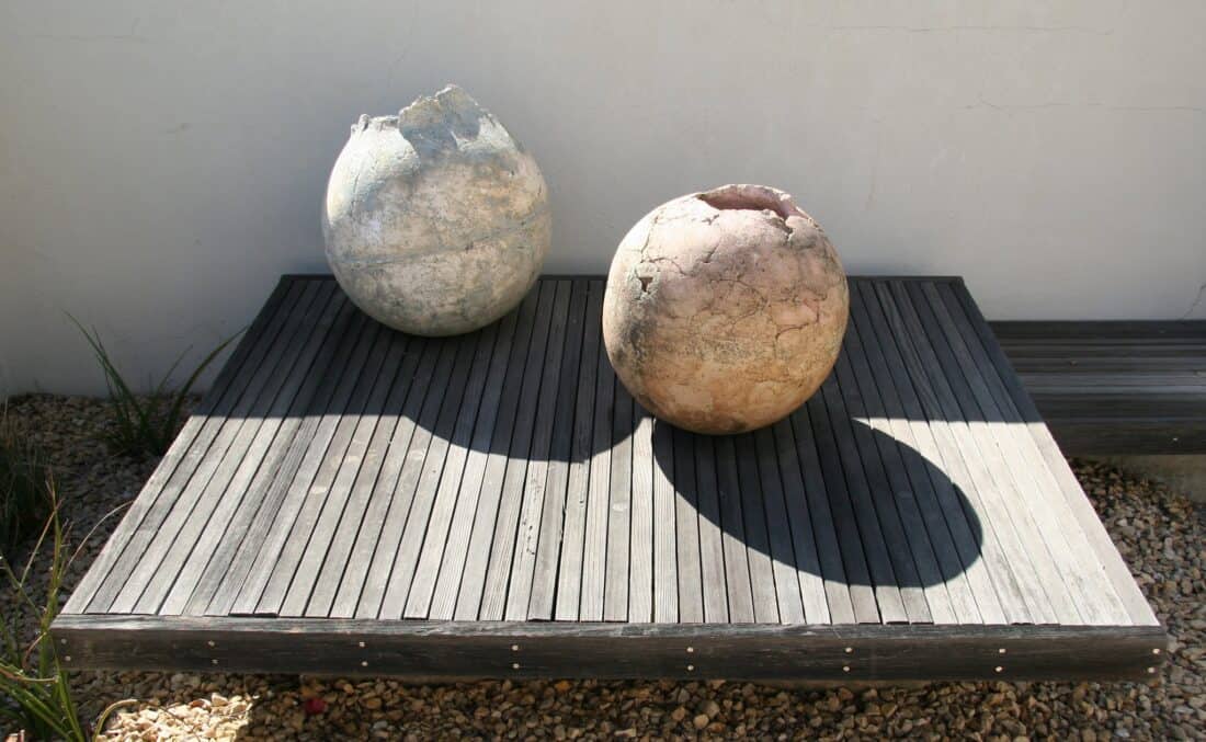 A photograph of two weathered, spherical clay pots displayed on a wooden platform in the Daily Garden. The pots, one with a light-gray hue and the other a light-brown hue, cast distinct shadows on the platform. The background features a minimalist white wall and gravel ground in Santa Barbara.