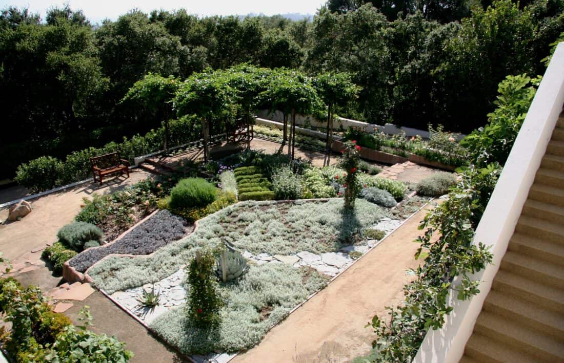 An aerial view of Isabelle Greene's well-maintained Santa Barbara garden features neatly trimmed bushes, various types of plants, and a gravel pathway. A wooden bench sits under a shaded pergola with leafy vines. A staircase descends alongside the garden, bordered by flowers and greenery.