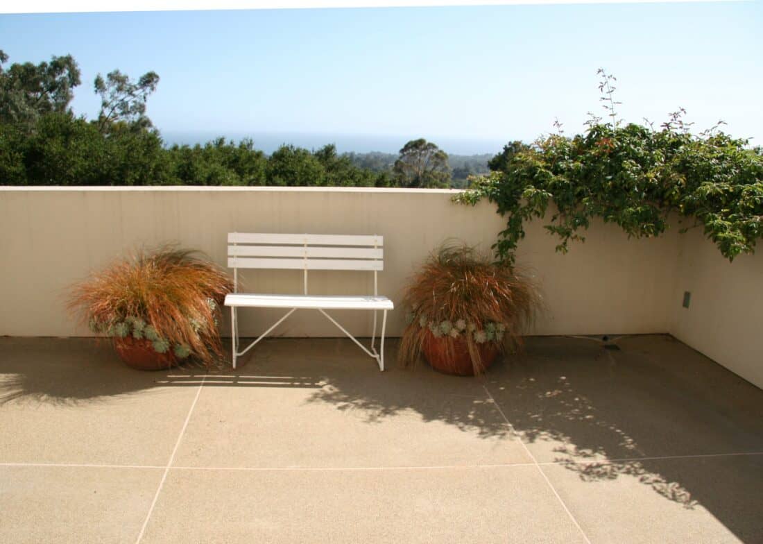 A minimalist outdoor patio with a white bench situated between two potted plants in a design reminiscent of Isabelle Greene’s work. A low wall behind the bench is adorned with lush green ivy. In the background, trees and a distant, hazy horizon are visible under Santa Barbara's clear sky.