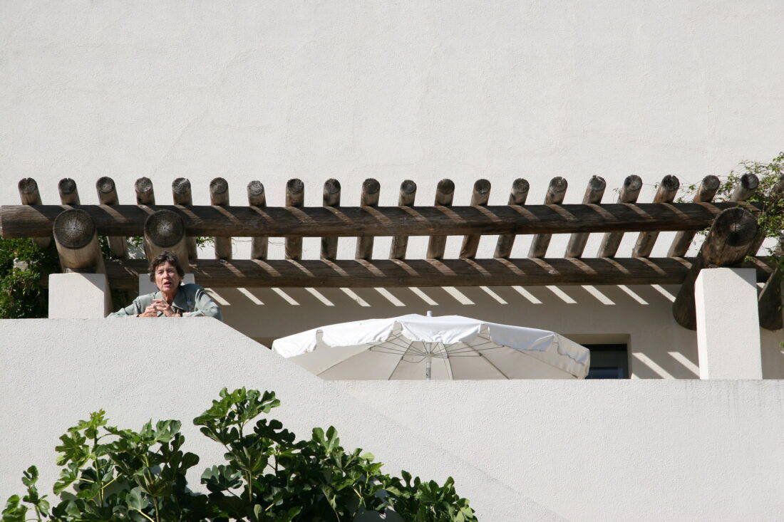 A person is standing on a terrace with a wooden pergola above and a white umbrella below. The background is a plain, light-colored wall, and leafy green plants are visible in the foreground, evoking the serene designs of Isabelle Greene. The person appears to be looking into the distance.