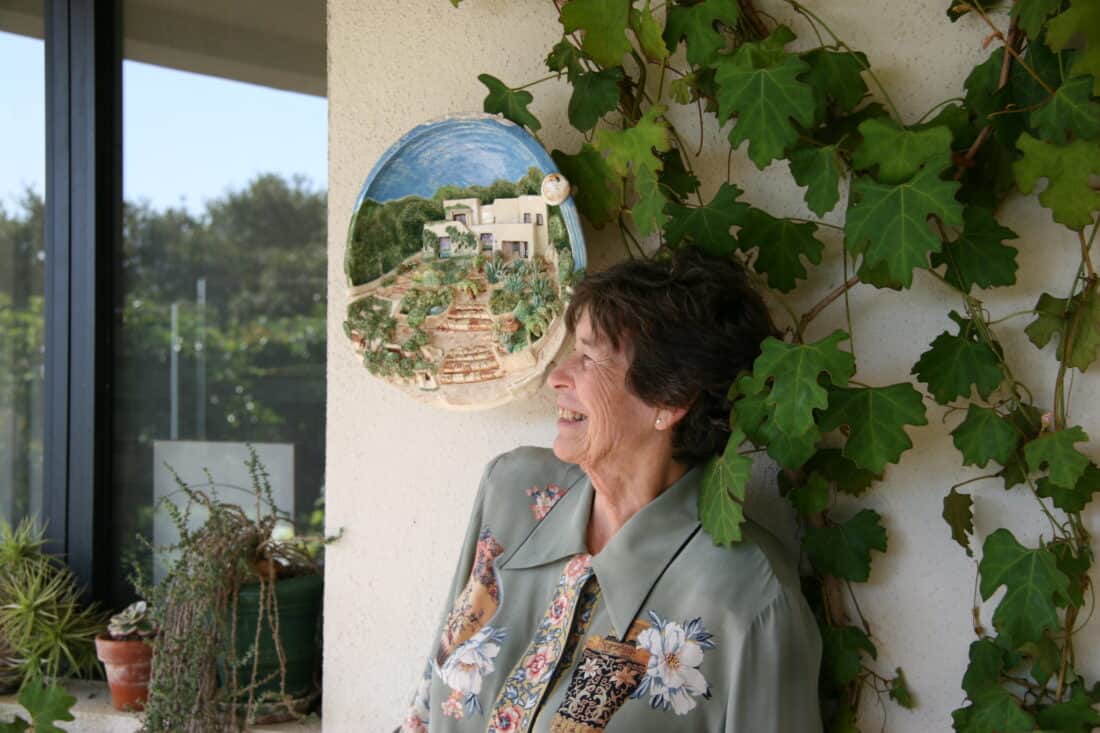 An elderly woman with short, graying hair stands smiling and leaning against a wall decorated with green ivy and a round ceramic art piece depicting a house and garden. She wears a floral-patterned blouse reminiscent of Isabelle Greene's designs. Potted plants and a large window nearby echo the charm of Daily Garden in Santa Barbara.