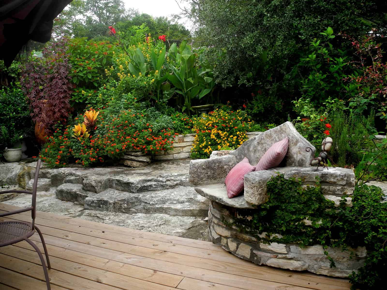 A peaceful garden scene with a stone bench adorned with pink cushions, surrounded by lush greenery and vibrant flowers. The foreground features a wooden deck and a metal chair, while stone steps lead up to the colorful garden bed—a perfect illustration of how to design garden layouts harmoniously.