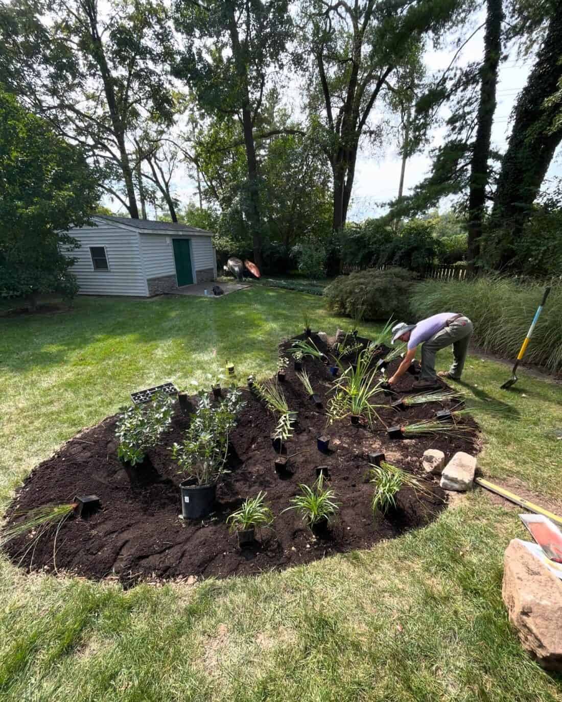 In the serene backyard, a person is gardening, creating a sustainable rain garden by arranging and planting small plants in a freshly prepared flower bed. A shed and various tools stand ready, surrounded by green grass and trees. This setup is perfect for mitigating mud while nurturing nature.