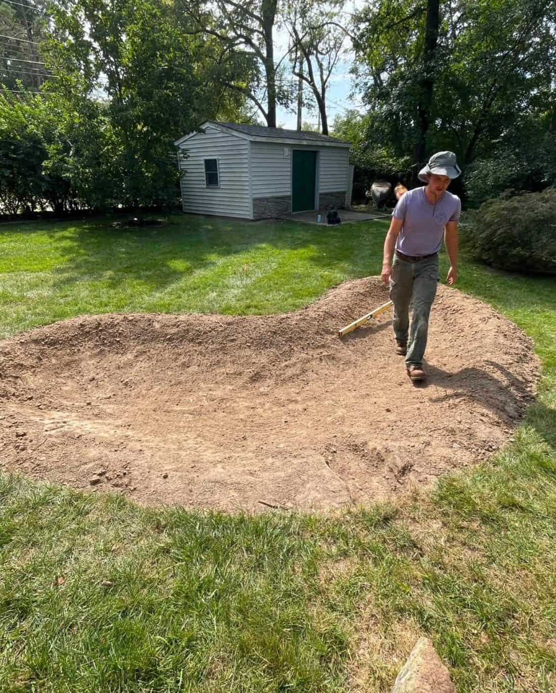 A person in a hat and purple shirt walks beside a dug-out dirt area in the grassy backyard, likely prepping for a rain garden. Behind them is a small white shed with a green door surrounded by trees, highlighting the sustainable landscaping efforts underway.