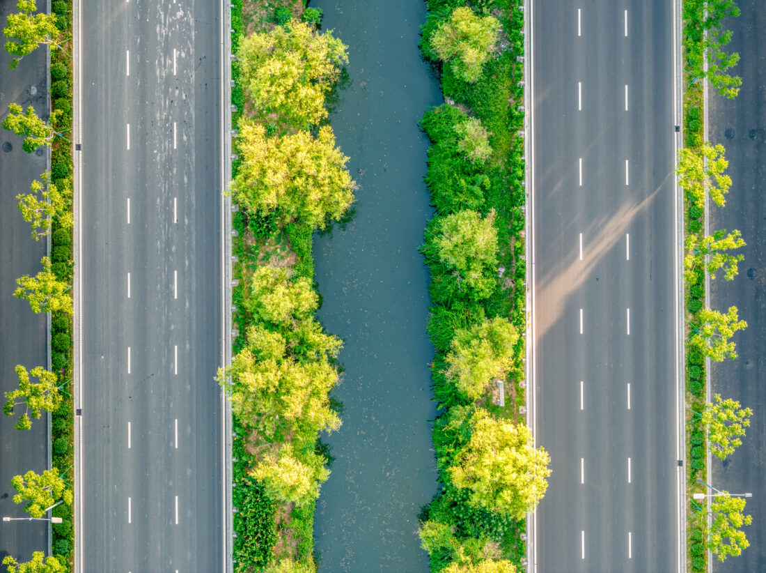 Aerial view of two parallel highways separated by a narrow river, with green trees and lush gardens lining both sides of the water and along the roads.